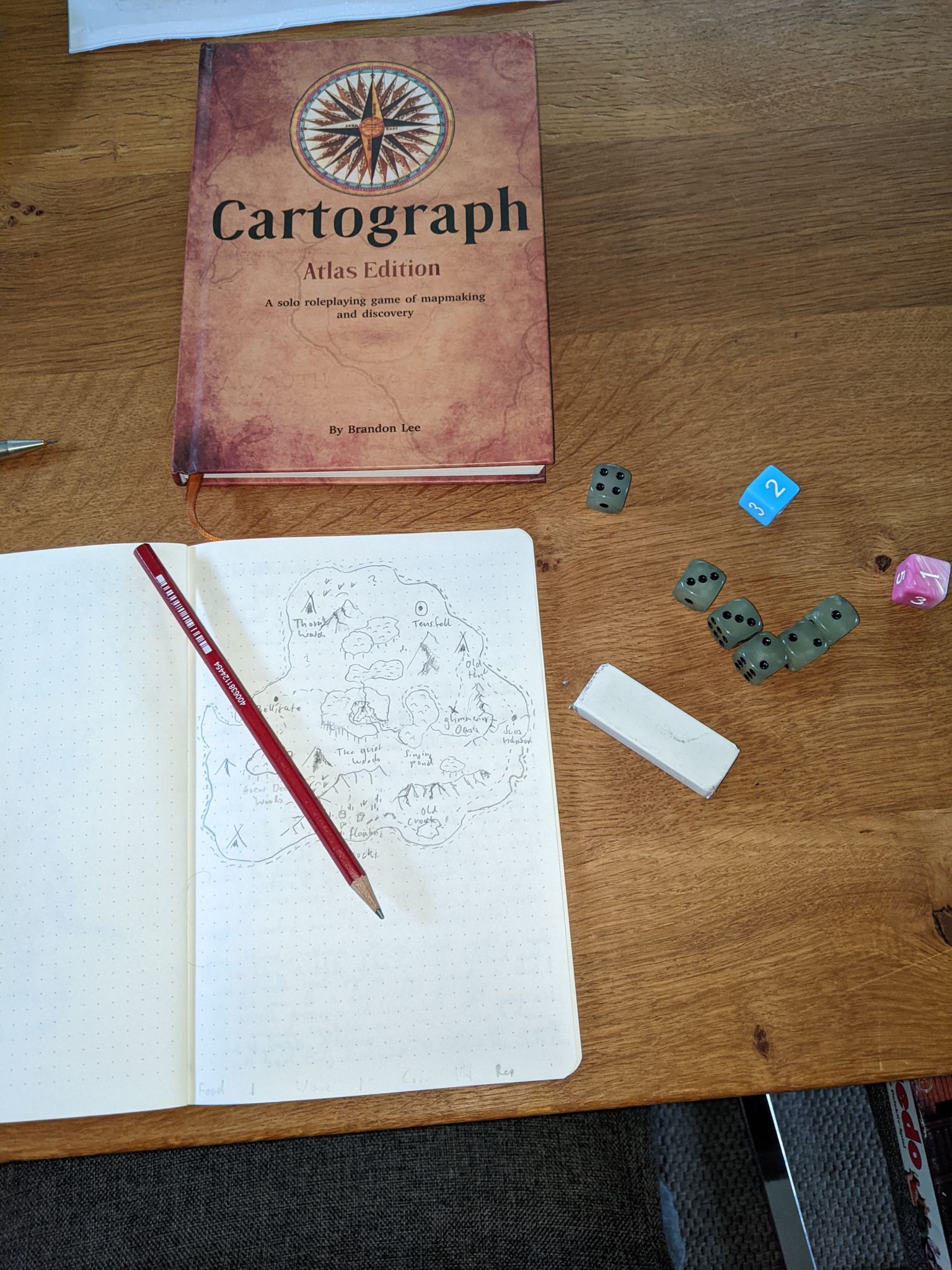 A notebook with a drawn island full of landmarks and place names. Above a closed hardcover book of the game Cartograph Atlas edition. On the side a pencil and some dice